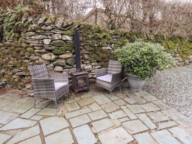 An outdoor seating area with two chairs a small stove and a potted plant against a mossy stone wall at Hart Head Barn in Rydal