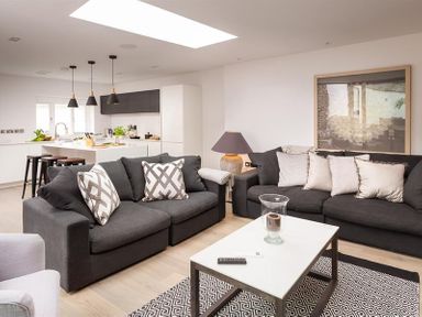 A living room with two dark gray sofas with pillows a white coffee table with remote a rug and a kitchen area with stools and pendant lights at Underhowe in Grasmere