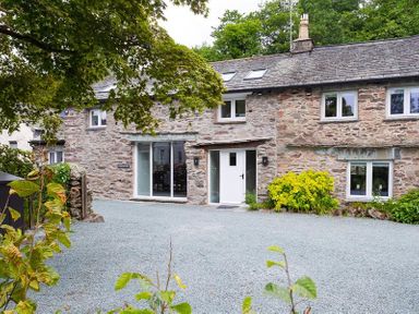 A stone house with a white door and windows surrounded by plants and trees at Underhowe in Grasmere