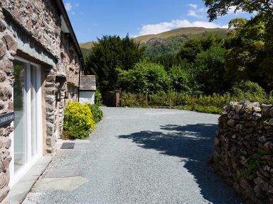 A stone building with a gravel driveway and green bushes with hills in the background at Underhowe Grasmere