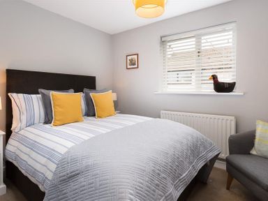 A bedroom with a bed with striped bedding and yellow pillows a window with blinds and a duck ornament a gray chair and a framed picture at Hill Cottage in Ambleside