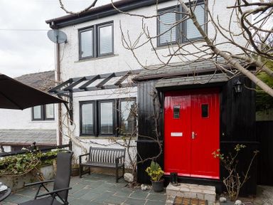 The front exterior of a house with a red double door a wooden bench potted plants and patio chairs at Hill Cottage in Ambleside