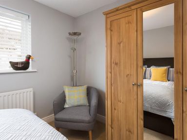 A bedroom corner with a grey armchair and cushion next to a wooden wardrobe with a mirrored door at Hill Cottage in Ambleside