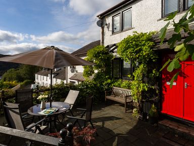 An outdoor patio with a round table and umbrella surrounded by chairs next to a house with a red door and climbing plants at Hill Cottage Ambleside
