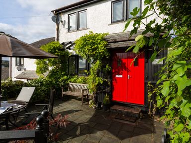 A patio with a table, chairs, an umbrella, a wooden bench, plants, and a house with a red door at Hill Cottage in Ambleside