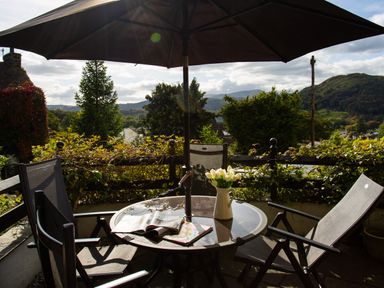A patio table with an umbrella three chairs a vase with flowers and magazines on the table overlooking a garden and hills at Hill Cottage in Ambleside