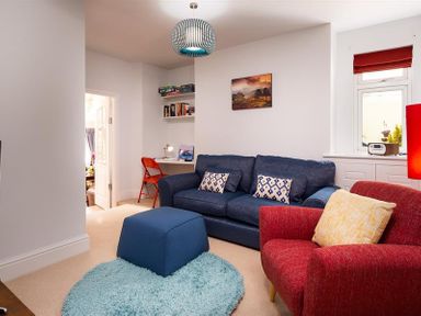 A living room with a blue sofa and matching ottoman a red armchair a small desk with a red chair and shelves a window with red blinds and a landscape painting on the wall at Ellerthwaite House in Windermere