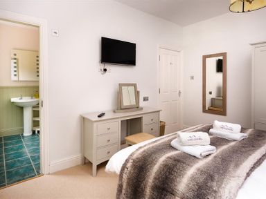 A bedroom with a bed covered with a fur blanket towels on the bed a desk with a mirror a wall-mounted television a door leading to a bathroom and a wall mirror at Ellerthwaite House in Windermere