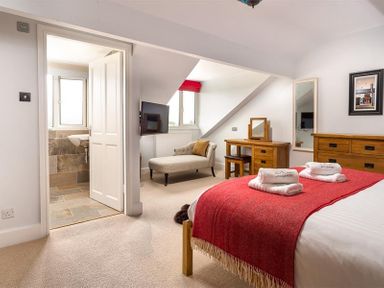 A bedroom with a bed covered by a red blanket towels on the bed a wooden dresser and vanity chair near a window and an open door leading to a bathroom at Ellerthwaite House in Windermere