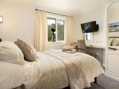 A bedroom with a bed, pillows, folded towels, window with curtains, desk with chair, wall-mounted TV and mirror at Heathfield in Keswick