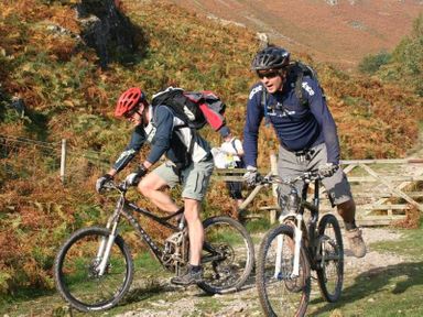 Two people riding mountain bikes on a dirt path with ferns and hills in the background at Heathfield in Keswick