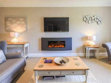 A living room with a wall mounted TV and fireplace two side tables with lamps and a coffee table with a bowl and remote controls at Pure Grace in Ambleside