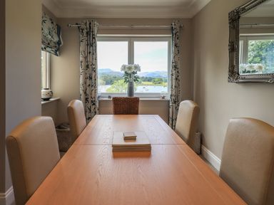A dining room with a wooden table beige chairs a window with floral curtains and a view of a lake and hills at Pure Grace in Ambleside