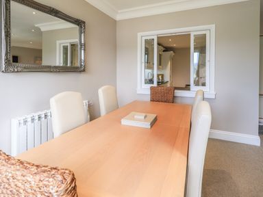A dining room with a wooden table surrounded by light-colored chairs and a wicker chair with a window showing a kitchen at Pure Grace in Ambleside