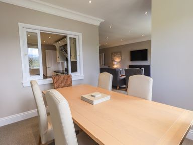A dining table with chairs near a window opening to a kitchen and a living room with armchairs and a wall-mounted television at Pure Grace in Ambleside