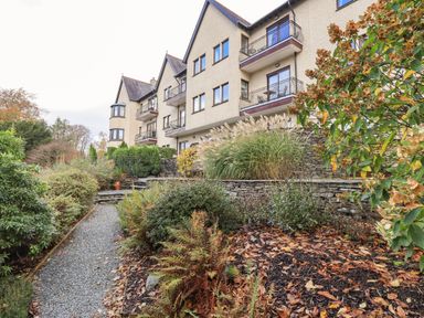 A gravel pathway surrounded by plants and shrubs leading to a beige multi-story building with balconies and a stone retaining wall at Pure Grace in Ambleside