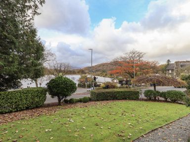 A garden with grass and bushes near a road with trees and water beyond at Pure Grace Ambleside