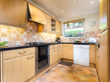 A kitchen with wooden cabinets beige tiled floor dishwasher oven and a window showing an outside view at Honister Cottage Ambleside