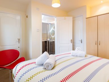 A bedroom with a bed covered by a striped duvet and rolled towels on top two red chairs a wooden wardrobe and an open door leading to a bathroom at Honister Cottage in Ambleside