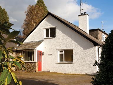 A white cottage with a red door and stone accents surrounded by trees at Honister Cottage in Ambleside