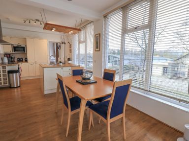 A dining area with a wooden table and four blue chairs next to large windows and a kitchen area at Windows Ambleside