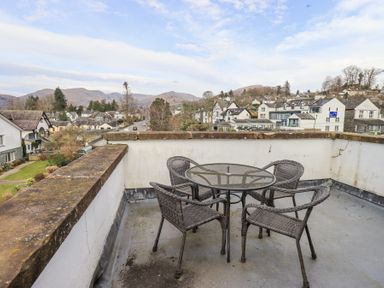 A rooftop patio with a glass round table and four wicker chairs overlooking houses and hills at Windows in Ambleside