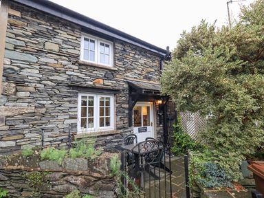 The exterior of a stone cottage with a white door and windows a small patio with a table and chairs and surrounding greenery at Cobblestone Cottage in Ambleside
