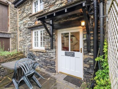 A stone cottage entrance with a white door and plastic chairs stacked on a table at Cobblestone Cottage in Ambleside