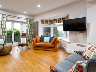 A living room with an orange sofa blue and patterned cushions a grey armchair patterned curtains and a wall-mounted TV at Wynford in Ambleside