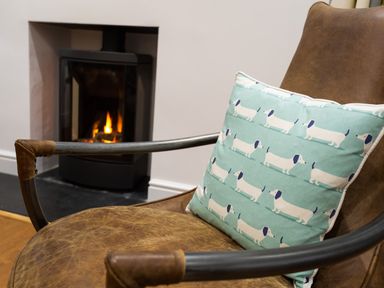 A brown leather chair with a cushion featuring white dog illustrations in front of a lit fireplace at Wynford in Ambleside