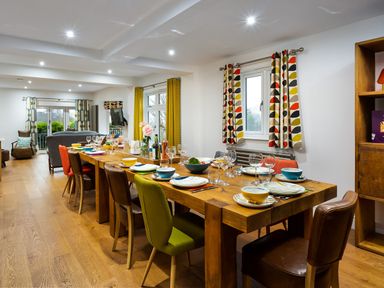 A dining room with a long wooden table set with plates bowls and glasses surrounded by mixed chairs and windows with patterned curtains at Wynford in Ambleside