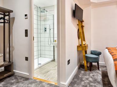 A bedroom with a wooden ladder holding a tv and clock a blue chair a bed with an orange blanket and an open door showing a tiled shower at Wynford in Ambleside
