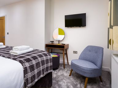 A bedroom with a bed covered with a checkered blanket and towels on top a wooden desk with a round mirror and a stool a blue chair mounted TV on the wall and curtains at Wynford in Ambleside