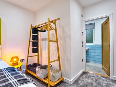 A bedroom corner with a wooden clothes rack holding hangers and baskets next to a yellow nightstand with an orange lamp at Wynford in Ambleside