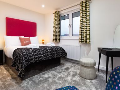 A bedroom with a bed with a red headboard and patterned blanket a window with patterned curtains a small black dressing table and a round stool at Wynford in Ambleside