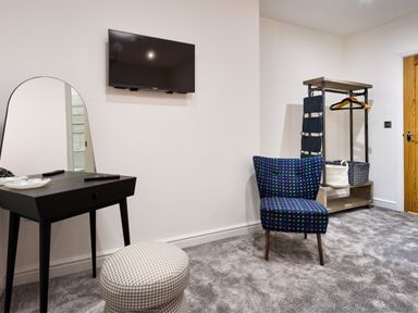 A room with a wall-mounted TV a black dressing table with mirror a patterned ottoman a blue chair with multicolored dots and an open closet with baskets at Wynford in Ambleside