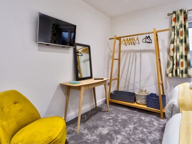 A room with a wall-mounted TV wooden desk with a mirror wooden clothes rack with hangers storage baskets and a yellow chair at Wynford in Ambleside