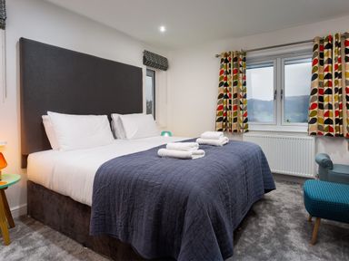A bedroom with a large bed with white pillows and a dark gray quilt two windows with patterned curtains and folded towels on the bed at Wynford in Ambleside