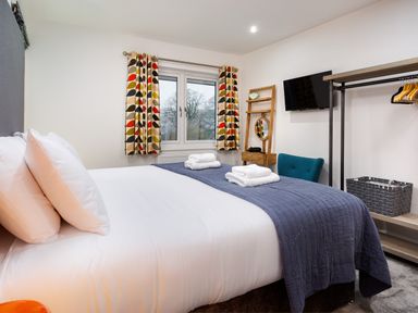 A bedroom with a bed with white sheets and blue coverlet next to a window with curtains a wall-mounted TV a blue chair a wooden dressing table with mirror and an open wardrobe with hangers and basket at Wynford Ambleside
