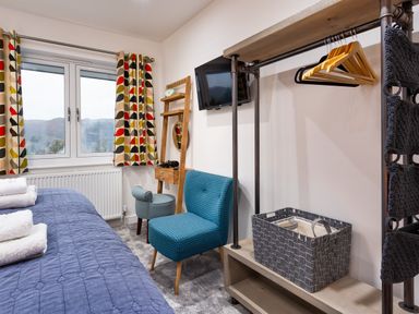 A bedroom with a blue bedspread and folded towels a window with patterned curtains a small chair a wall-mounted tv a wooden rack with hangers and a basket at Wynford Ambleside