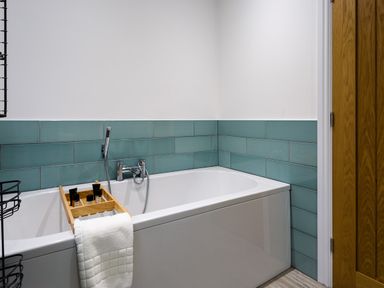 A bathroom with a white bathtub a wooden bath tray with toiletries a white towel teal wall tiles and a wooden door at Wynford in Ambleside