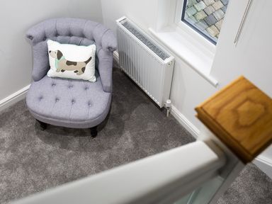 A gray upholstered chair with a dog cushion next to a white radiator under a window in a carpeted corner space at Wynford in Ambleside