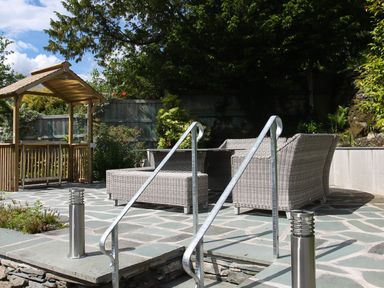 An outdoor patio with woven seating furniture a wooden pergola metal handrails and stone flooring at Wynford in Ambleside