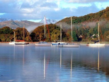 A lake with several sailboats moored near a wooded shoreline with hills in the background at Wynford in Ambleside
