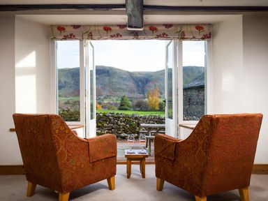 A room with two armchairs and a small table with glasses of wine facing open doors showing hills and a stone wall at Horrockwood Farm in Watermillock