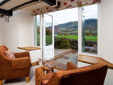 A seating area with two armchairs and a small table with two glasses next to open doors showing a stone patio and countryside at Horrockwood Farm in Watermillock