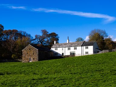 A white farmhouse and stone barn on a grassy hill with trees in the background at Horrockwood Farm in Watermillock