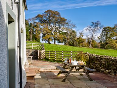 An outdoor patio with a wooden picnic table set with food and drinks next to a white building and fenced green field at Horrockwood Farm in Watermillock