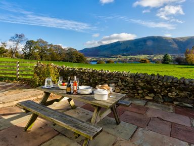 A wooden picnic table with bottles and dishes on a stone patio overlooking a green field and hills at Horrockwood Farm in Watermillock