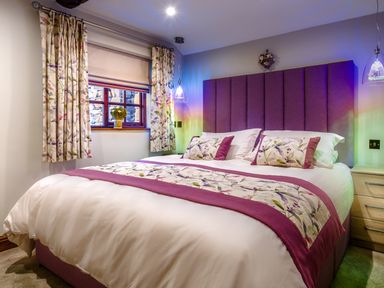 A bedroom with a purple headboard a bed with floral and purple bedding floral curtains and a window with a flower vase at Tipsy Gin Cottage in Crosthwaite